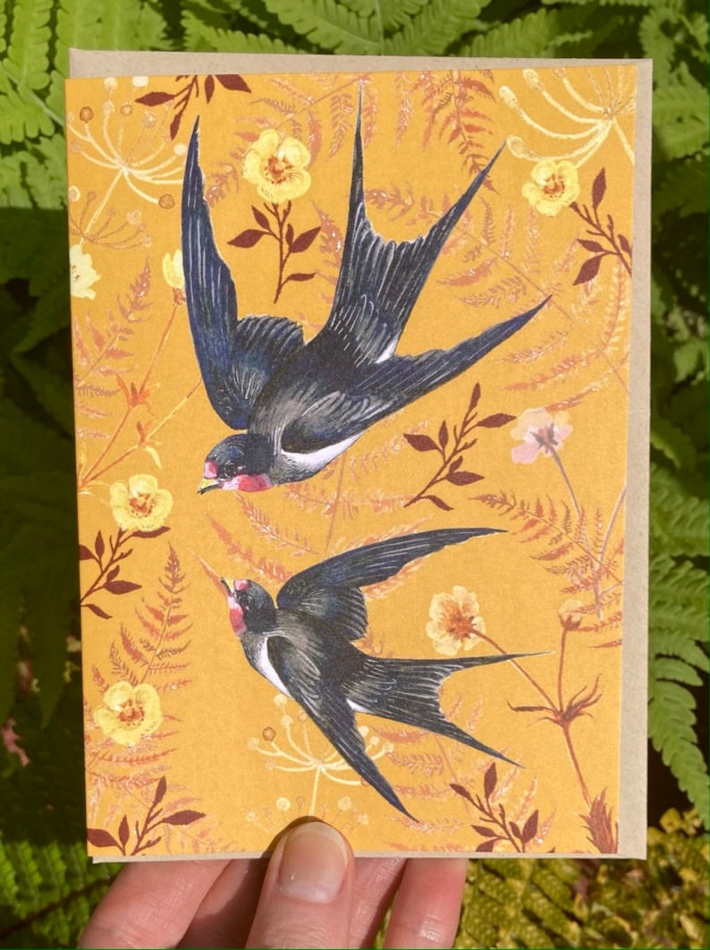 May include: A greeting card with a yellow background featuring two black and white swallows in flight. The background is filled with a floral pattern of yellow flowers and brown leaves.