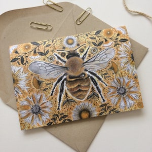 May include: A greeting card featuring a detailed illustration of a bee with outstretched wings, surrounded by yellow and white flowers and foliage. The card is set on a brown envelope, with gold paper clips and twine.