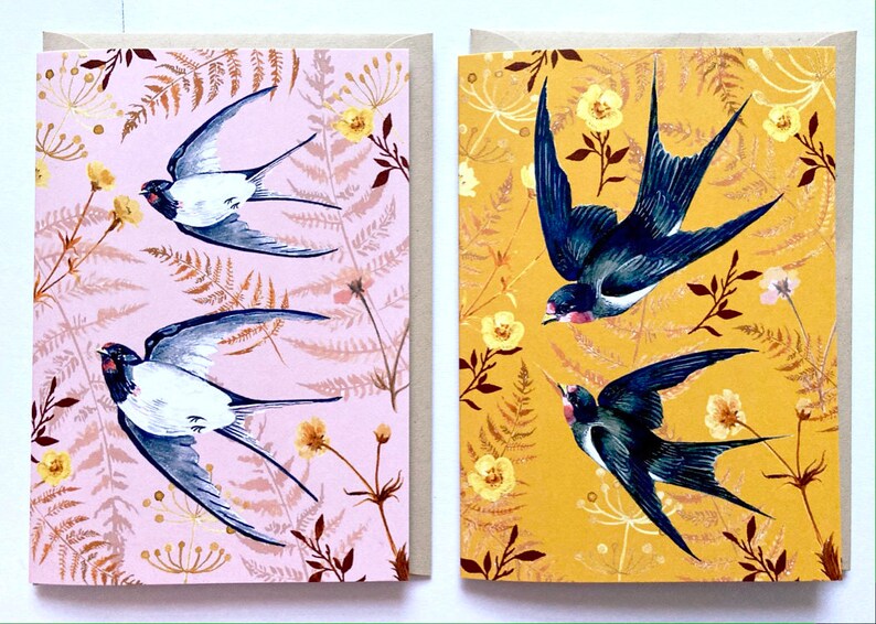 May include: Two greeting cards with a floral pattern and swallows in flight. The card on the left has a pink background with brown and white flowers and ferns. The card on the right has a yellow background with yellow and white flowers and ferns.
