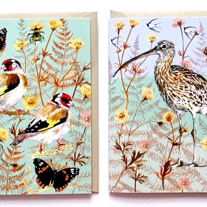 May include: Two greeting cards with nature-themed illustrations. The first card features two goldfinches perched on branches with a butterfly and a beetle. The second card features a long-billed curlew standing in a field of flowers.