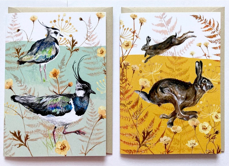 May include: Two greeting cards with nature-themed illustrations. The card on the left features a pair of lapwings in a green and brown floral pattern. The card on the right features a pair of hares in a yellow and brown floral pattern.