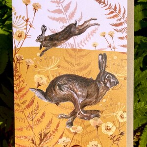 May include: A greeting card with a brown background featuring two brown rabbits hopping through a field of yellow flowers and brown ferns.