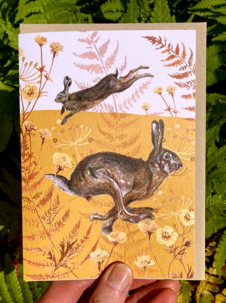 May include: A greeting card with a brown background featuring two brown rabbits hopping through a field of yellow flowers and brown ferns.