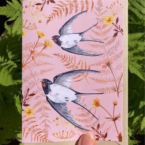 May include: A pink card with a floral pattern featuring two gray and white birds in flight. The birds have red accents on their beaks and are flying against a pink background with brown and yellow floral accents.
