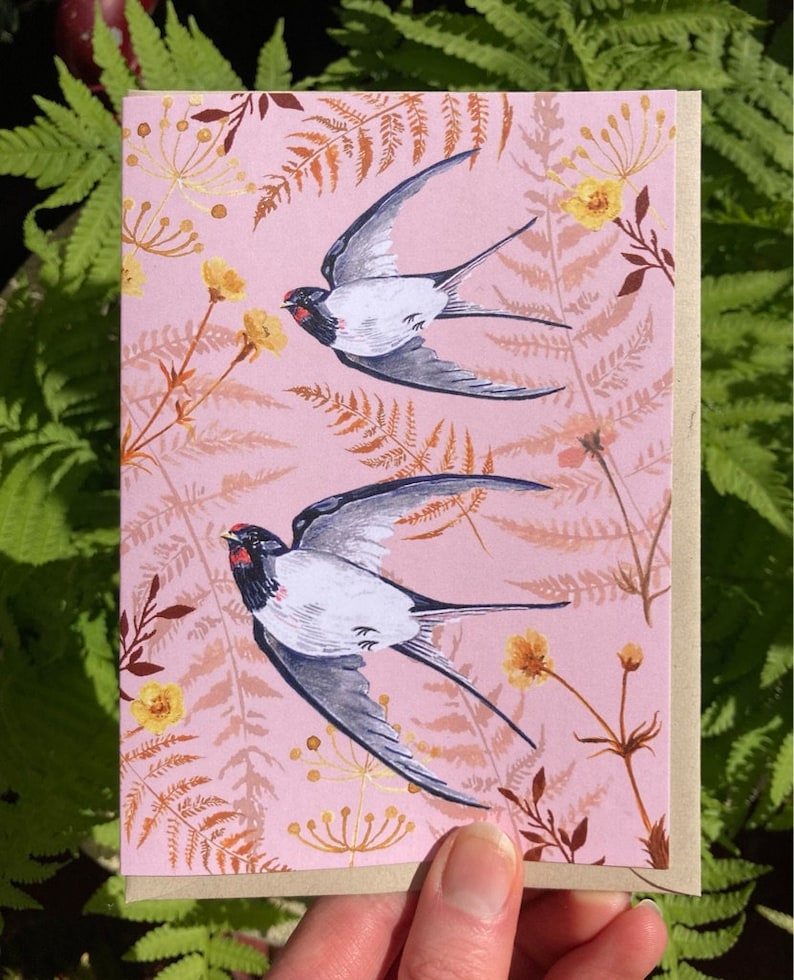 May include: A pink card with a floral pattern featuring two gray and white birds in flight. The birds have red accents on their beaks and are flying against a pink background with brown and yellow floral accents.