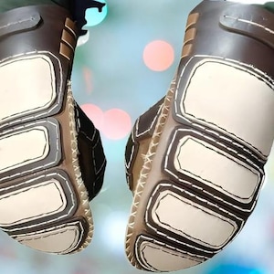 May include: A pair of brown and cream-coloured gloves featuring a segmented design with padded rectangular panels on the back of the hand. The gloves are constructed with visible white stitching along the edges of each panel and a strap at the wrist.