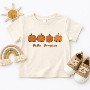 May include: A cream-colored short-sleeve t-shirt featuring a graphic of four orange pumpkins with faces and the text "HELLO PUMPKIN" printed in brown letters. The shirt is displayed alongside a crocheted sun, a rainbow, and two tan sneakers.