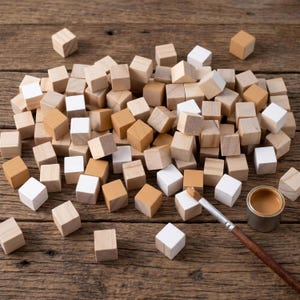 May include: A collection of small wooden cubes in natural, white, and tan finishes scattered across a surface. A paintbrush with a wooden handle and a small metal container filled with tan paint are positioned near the wooden blocks.