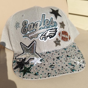 Könnte beinhalten: Eine hellgraue Baseballkappe mit dem Logo und Schriftzug der Philadelphia Eagles, verziert mit verschiedenen sternförmigen Aufnähern, einem footballförmigen Aufnäher und zahlreichen kleinen, funkelnden Strasssteinen in Silber, Petrol und Schwarz.