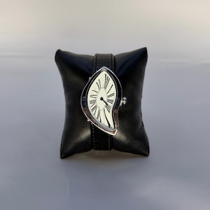 May include: A wristwatch with an asymmetrical, teardrop-shaped silver case and a white dial featuring elongated black Roman numerals. The watch has a black leather strap with visible stitching and is mounted on a black square display cushion.