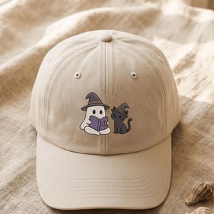 Könnte beinhalten: Eine beige Baseballkappe mit einer Stickerei, die einen weißen Geist und eine schwarze Katze zeigt, beide tragen spitze Hexenhüte. Der Geist hält ein offenes violettes Buch mit einem Mondsichel-Symbol auf dem Einband. Die Kappe hat eine feste Form.