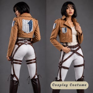 May include: A tan cropped jacket with a blue and white wing emblem on the back and shoulders, paired with white trousers, a brown leather harness system with multiple straps and buckles, and dark brown boots. The outfit includes a white top.
