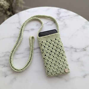 May include: A light sage green crochet phone pouch featuring a textured shell stitch pattern, a long matching shoulder strap, and a single silver-toned circular snap button closure at the bottom, containing a black rectangular smartphone.