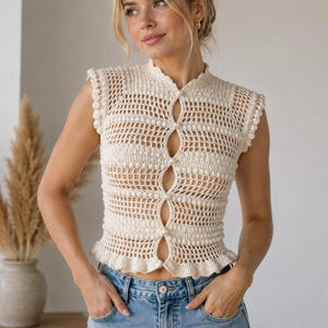 May include: A cream-colored sleeveless crochet top featuring a high neckline, a vertical row of small white pearl-like buttons down the center, and a ruffled hem. The garment is constructed with an open-work grid pattern and textured bobble details.