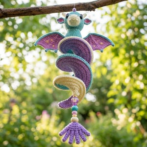 Op de afbeelding: Een gehaakte draak als hangende decoratie met een groenblauwe kop, kleine hoorns, paarse vleugels met gele sterrenborduursels en een spiraalvormig lichaam in paars en crème. De staart eindigt met een kralensnoer en een paarse gehaakte kwast.