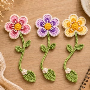 May include: Three handmade crochet flower bookmarks with long green stems and leaves. Each flower features five petals in pink, purple, or yellow, with a central yellow circle and small white bead accents. A small white flower with a pearl center sits at the base.