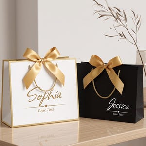 Könnte beinhalten: Eine weiße Geschenktüte mit Goldrand und goldener Schleife, beschriftet mit dem Namen Sophia und dem Text Your Text, neben einer schwarzen Geschenktüte mit Goldrand und goldener Schleife, beschriftet mit Jessica und dem Text Your Text.