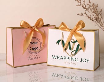 May include: Two rectangular gift bags with gold ribbon bows and twisted cord handles. The left bag is light pink with a circular logo and the text Your logo and Kirsten. The right bag is white with a green logo and the text WRAPPING JOY STUDIO.