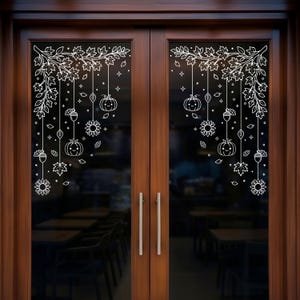 Puede incluir: Un conjunto de pegatinas blancas para ventanas aplicadas en puertas de cristal dobles, con un diseño otoñal simétrico de hojas de arce, girasoles, bellotas y calabazas sonrientes colgantes, con pequeñas estrellas y follaje.