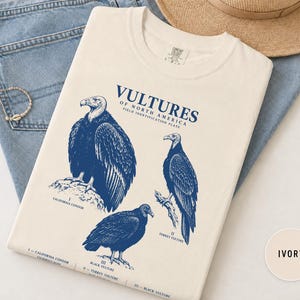 Puede incluir: Una camiseta color marfil con un estampado gráfico azul titulado VULTURES OF NORTH AMERICA FIELD IDENTIFICATION PLATE. El diseño muestra ilustraciones de un cóndor de California, un aura gallipavo y un zopilote negro con etiquetas.