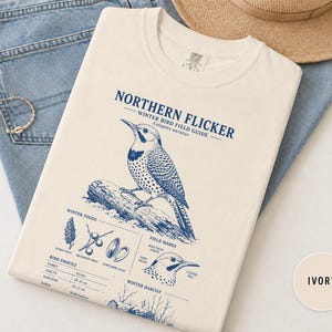 May include: An ivory t-shirt featuring a blue graphic print of a Northern Flicker bird. The design includes the text 'NORTHERN FLICKER WINTER BIRD FIELD GUIDE', illustrations of the bird, winter foods, and bird profile data with measurements in cm.