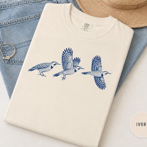May include: An ivory t-shirt featuring a dark blue graphic print of three birds in flight. The birds are depicted in a sequence showing one perched and two flying with detailed spotted patterns on their wings and bodies. The shirt has a crew neckline.