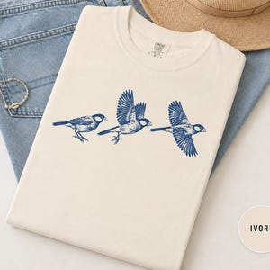 May include: An ivory-colored t-shirt featuring a blue graphic print of three birds in various stages of flight. The shirt has a crew neckline and a label at the inner collar. The design shows one bird perched and two birds with wings spread.