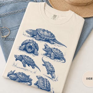 May include: An ivory t-shirt featuring a blue graphic print of multiple snapping turtles in various poses. The shirt has a crew neckline and a tag printed on the inside collar area. The design displays detailed illustrations of the turtles.