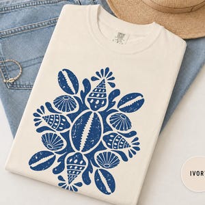 May include: An ivory short-sleeved t-shirt featuring a large, centred graphic print in dark blue. The design consists of a symmetrical arrangement of various seashells, including cowrie and scallop shells, rendered in a distressed, textured style.