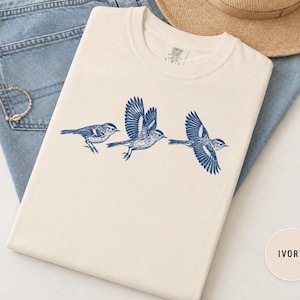 Puede incluir: Una camiseta de color marfil con un estampado gráfico azul de tres pájaros en varias posiciones de vuelo. La camiseta tiene cuello redondo y una etiqueta en el interior del cuello. El diseño muestra un pájaro posado y dos volando.