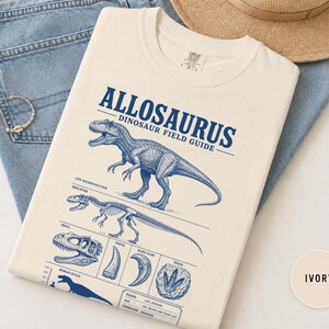 May include: An ivory t-shirt featuring a blue graphic print titled ALLOSAURUS DINOSAUR FIELD GUIDE. The design includes a detailed illustration of an Allosaurus, a skeleton, a skull, a tooth, a hand claw, a track, and a human scale comparison.