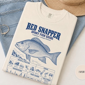 Peut inclure: Un t-shirt ivoire avec un motif bleu représentant un vivaneau rouge et le texte Red Snapper Sport Fish Guide Lutjanus campechanus. Sous le poisson se trouvent des tableaux intitulés Water & Structure, Presentations, Forage, Prime Seasons et Size & Power.