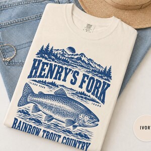 May include: An ivory t-shirt featuring a blue graphic print of a rainbow trout swimming in a river with mountains and pine trees in the background. The text reads Henry's Fork, Island Park, Idaho, and Rainbow Trout Country in a bold font.
