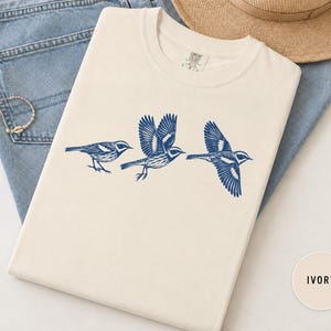 May include: An ivory t-shirt featuring a dark blue graphic print of three birds in various stages of flight. The birds are depicted in a detailed, etched style, showing wings spread and folded. The shirt has a crew neckline and a standard fit.