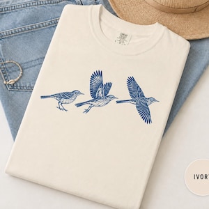May include: An ivory crew neck t-shirt featuring a blue graphic print of three birds in various stages of flight. The shirt has a tag at the neckline with the text Comfort Colors and the size L-G. The design shows detailed line work on the birds.