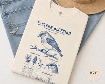 Puede incluir: Una camiseta de color marfil con un estampado gráfico azul de un azulejo oriental. El diseño incluye el texto Eastern Bluebird, Winter Bird Field Guide, Sialia sialis, Winter Foods, Field Marks y Winter Habitat, junto con ilustraciones de aves.