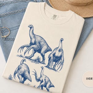 Op de afbeelding: Een ivoorkleurig t-shirt met een blauwe grafische print van vier therizinosaurus-dinosaurussen in verschillende houdingen. Het shirt heeft een ronde hals en een label aan de binnenkant van de kraag. Het ontwerp toont de dinosaurussen met lange klauwen.
