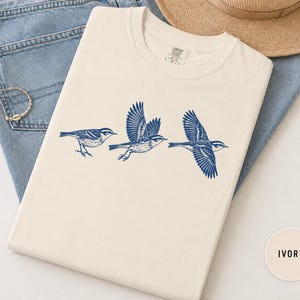 May include: An ivory short-sleeved t-shirt featuring a graphic print of three birds in varying stages of flight in dark blue ink. The shirt has a crew neckline and a label at the inner collar. The design shows birds with detailed wings.