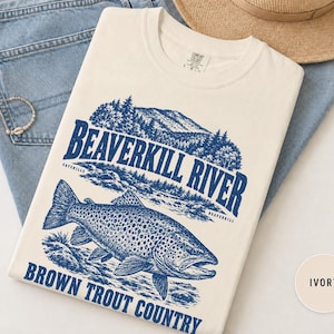 May include: An ivory short-sleeve t-shirt featuring a blue graphic print of a brown trout, a river, and mountains. The text reads BEAVERKILL RIVER, CATSKILLS, and BROWN TROUT COUNTRY. The shirt has a crew neckline and a tag indicating size L-G-G.