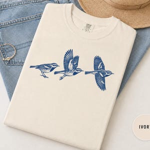 May include: An ivory crew neck t-shirt featuring a dark blue graphic print of three birds in various stages of flight. The shirt is made of cotton and displays a label at the inner neckline. The design shows the birds in a horizontal sequence.