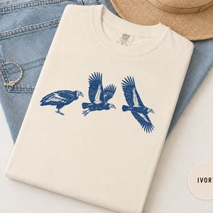 Puede incluir: Una camiseta de cuello redondo color marfil con un estampado gráfico azul oscuro de tres buitres en diferentes posiciones de vuelo y aterrizaje. La prenda tiene un diseño estándar de manga corta y una etiqueta de marca visible en el cuello.