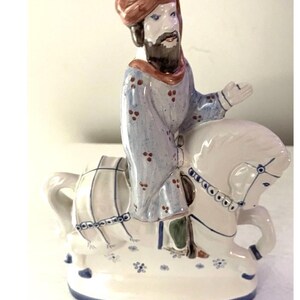 May include: A ceramic figurine of a person wearing a brown turban and a light blue patterned robe, riding a white horse with blue line details. The horse is positioned on a white base decorated with small blue floral accents and a blue trim.