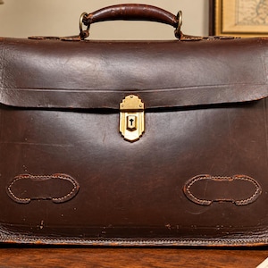 May include: A dark brown leather briefcase featuring a top handle, a rectangular flap, and a central gold-toned metal latch with a keyhole. The front of the bag displays two decorative stitched leather patches near the bottom edge.