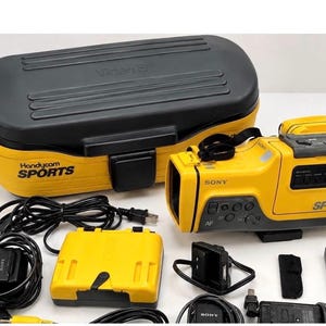 Peut inclure: Un caméscope Sony Handycam Sports jaune et noir avec une mallette de transport assortie jaune et noire marquée Video 8 et Handycam Sports. L'ensemble comprend une batterie jaune, des câbles d'alimentation noirs et divers accessoires électroniques.