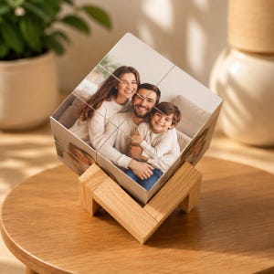 May include: A square photo cube puzzle featuring a family portrait of two adults and a child, divided into nine smaller square segments. The cube is supported by a light-colored wooden stand with a V-shaped base, displaying the image clearly.