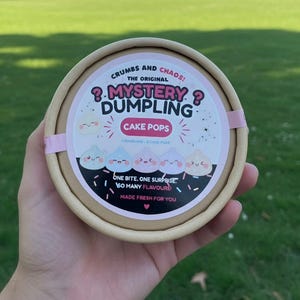 May include: A round cardboard container with a label reading Crumbs and Chaos! The Original Mystery Dumpling Cake Pops. The label features five colourful cartoon dumplings with faces, text stating 1 Dumpling = 9 Cake Pops, and One Bite. One Surprise. So Many Flavours! Made Fresh For You.