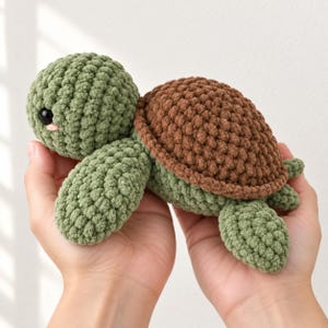 May include: A small, handmade crochet turtle plush toy featuring a textured sage green body and a rounded brown shell. The turtle has two large, glossy black eyes and small pink stitched details on its cheeks, crafted from soft yarn.