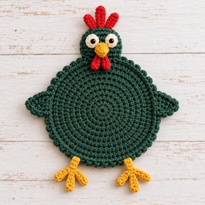 May include: A round, dark green crochet coaster shaped like a chicken. It features a red comb on top, a yellow beak, red wattle, white eyes with black pupils, and two yellow feet extending from the bottom. The texture is made of yarn stitches.
