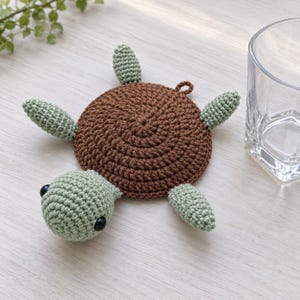 Puede incluir: Un posavasos de ganchillo con forma de tortuga, que presenta un cuerpo circular de color marrón oscuro y una cabeza de color verde claro con dos ojos de plástico negro. Tiene cuatro aletas de ganchillo verde claro en los lados del caparazón.