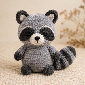 May include: A crochet raccoon plush toy featuring a light gray body, dark gray eye patches, and a white muzzle with a black nose. The raccoon has rounded ears with white centers and a striped tail with alternating light and dark gray bands.